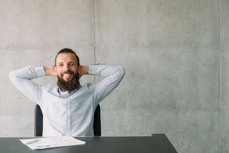 Successful professional career. Happy hipster guy sitting confidently at office desk, smiling. Gray wall background. Copy space.の写真素材