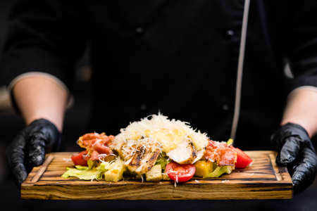 Italian restaurant serving. Chef holding Caesar salad with smoked chicken fillet, bacon chips on wooden board.の写真素材