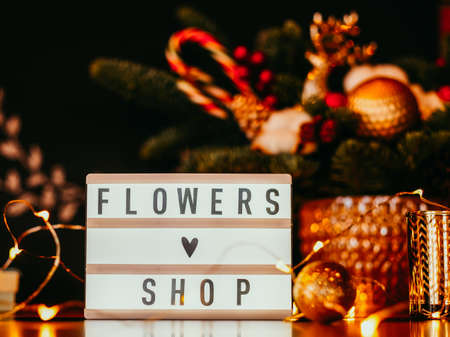 Handmade gift delivery. Flowers shop sign plate and fairy lights over blur fir tree arrangement.