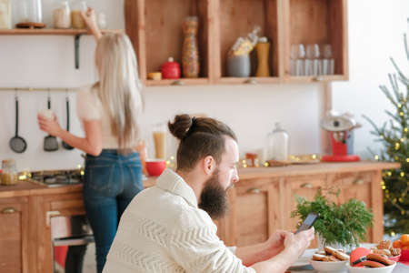 Christmas morning. Lady making breakfast in modern kitchen. Guy sitting at table, using smartphone.