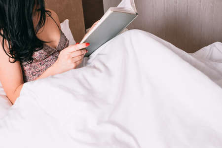 Home weekend leisure and hobby. Cropped shot of brunette woman in nightgown reading book in bed.の写真素材