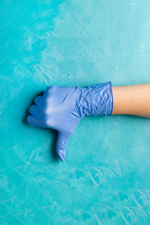 Bad protection. Virus outbreak. Hand in medicine gloves showing dislike gesture on turquoise blue background. Pandemic measures. COVID-19 preventionの写真素材