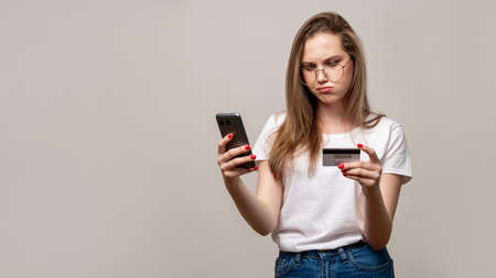 Bank fraud. Mobile scam. Confused woman in glasses using phone credit card isolated on gray copy...