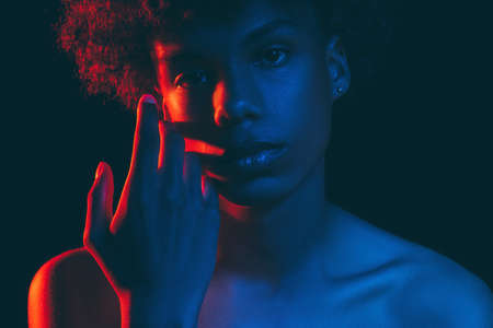 African man silhouette. Neon light portrait. Relaxed black guy in blue red glow touching face with hand looking at camera isolated on dark. Fashion model. Aesthetic cosmetology. Masculine powerの写真素材