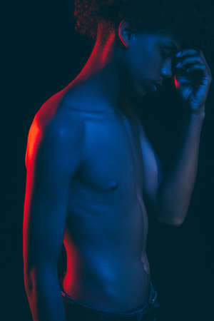 African man portrait. Neon light.Tired black male in blue red glow with shirtless torso touching forehead isolated on dark. Stress anxiety. Human tolerance. Ethnic problems. No racismの写真素材