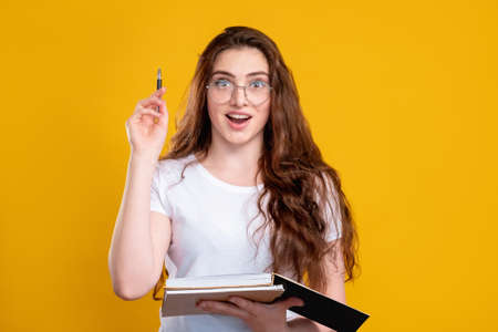 Inspired woman portrait. Creative idea. Enthusiastic female teacher in glasses standing with open notebook pointing up with pen isolated on yellow copy space background. Problem solution. Aha moment.の写真素材