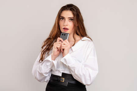 Scared woman portrait. Mobile scam. Worried lady with open mouth holding phone isolated on light...