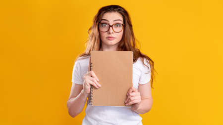 Scared student portrait. Exam anxiety. Worried woman in glasses with beige blank notebook isolated on orange copy space background. Panic attack. Higher education.の写真素材
