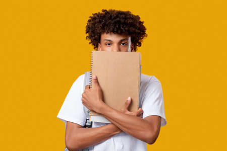Black teenager. Exam worry. Confused young male student hiding under notebook isolated on orange...