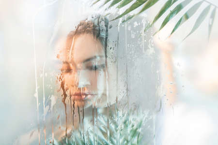 Spa therapy. Natural skincare. Double exposure of sensual peaceful woman face silhouette with exotic green leaves behind wet steamed glass with holographic iridescent blur glow. Body care freshness.の写真素材