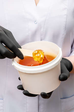 Honey depilation. Natural hair removal. Sugar wax in pot in female specialist hands wearing black latex gloves over blur white background. Professional cosmetology. Beauty procedure.の写真素材