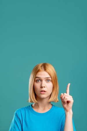 Overwhelmed woman. Exciting opportunity. Portrait of inspired blonde lady in blue t-shirt pointing up at copy space isolated on teal commercial background. Surprising idea.の写真素材