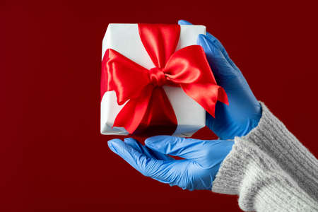 Happy New Year 2021. Stay safe. Female hands in medicine gloves holding present box taped ruby...