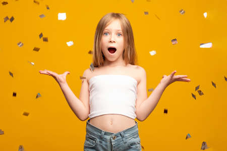 Holiday surprise. Kid party celebration. Festive fun. Young winner portrait. Amazed birthday girl in confetti rain isolated on orange background.の写真素材