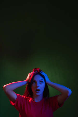 Shocked woman portrait. Special offer. Unexpected news. Amazing opportunity. Overwhelmed lady in red blue neon light looking up at copy space isolated on dark night green advertising background.の写真素材