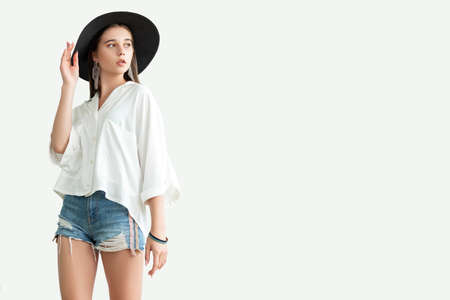 Summer outfit. Trend stylish look. Young woman in white shirt jeans shorts and wide-brimmed black hat looking copy space isolated on neutral. Casual eleganc. Advertising backgroundの写真素材