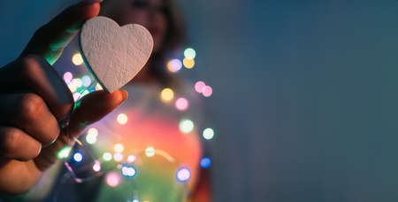 Love symbol. Romantic background. Festive decoration. Woman holding ceramic heart with fingers in colorful neon bokeh lights blur rainbow glow isolated on navy blue copy space.の写真素材