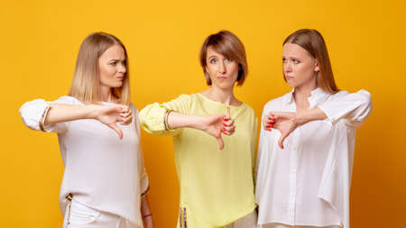 Group portrait of three dissatisfied ladies in white yellow blouse disapproving offer with thumbs down isolated on orange background.の写真素材