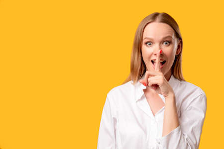 Portrait of enthusiastic amazed surprised woman in white shirt showing shhh with finger at open mouth isolated on orange copy space background.の写真素材