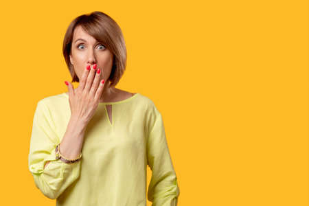 Shocked woman. Special offer. Omg impossible. Sale discount. Portrait of overwhelmed mature lady in yellow blouse looking at camera covering mouth isolated on orange copy space commercial background.の写真素材