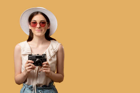 Holiday trip. Capture moments. Summer memories. Smiling woman sunglasses hat holding retro camera ready taking photo looking straight isolated on beige copy space. Advertising background.の写真素材