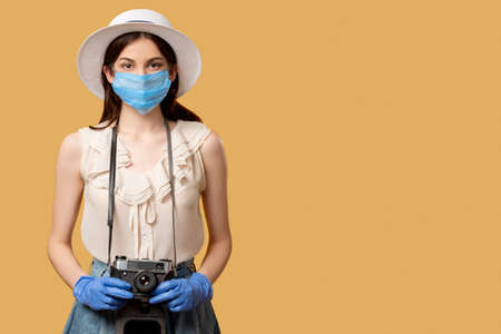 Holiday trip. Pandemic restriction. Travel photo. Summer memories. Woman in protective mask gloves holding retro camera isolated on beige copy space. Advertising background.の写真素材