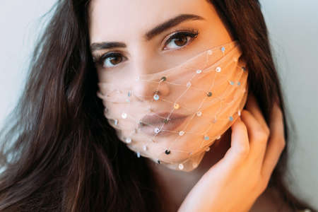 Pandemic beauty. DIY accessory. Fashion trend 2021. Portrait of sensual brunette woman with party makeup in glamour shiny golden mesh face mask looking at camera isolated on light neutral background.の写真素材
