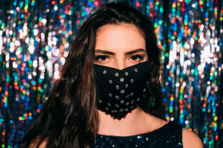 Pandemic fashion. Handmade accessory. Festive look. Portrait of brunette woman in black gothic style fabric protective face mask with rhinestone decor on blur colorful shiny tinsel background.の写真素材