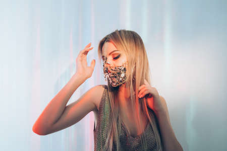 Pandemic party. Neon light portrait. Spiritual dance. Glamour woman in chain gem face mask matching gold dress enjoying trance in blue pink iridescent glow on copy space background.の写真素材