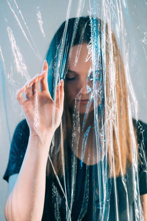 Pandemic self isolation. Quarantine loneliness. Covid-19 lockdown. Art portrait of blur tired trapped woman in black with closed eyes touching transparent polyethylene plastic film on blue background.の写真素材