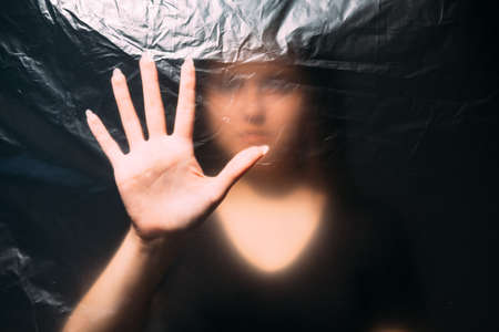 Covid-19 self isolation. Defocused silhouette. Quarantine loneliness. Woman hand touching creased texture clear polyethylene plastic film alone in darkness on black out of focus.の写真素材