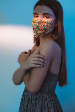 Quarantine fashion. Party look. Pandemic accessory. Covid-19 trend 2021. Tender woman in glamour gold chain face mask matching sparkling dress on blur blue white color gradient background.の写真素材