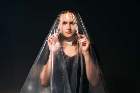 Art portrait. Plastic pollution. Environmental protection. Waste reuse. Blur pensive worried woman in transparent polyethylene film bag isolated on black copy space background.の写真素材