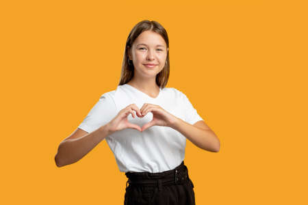 Love sign. Romantic message. Affection sympathy. Admiration compassion. Friendly cheerful teen girl in white showing heart gesture smiling isolated on orange empty space background.の写真素材