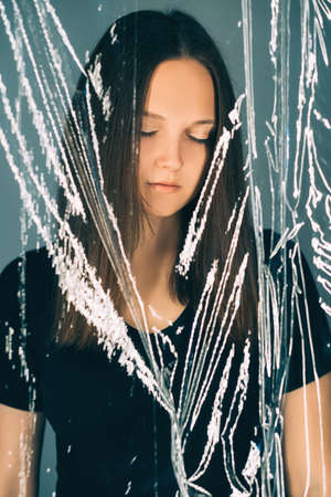 Female beauty. Detox treatment. Skincare wellness. Portrait of peaceful woman face with natural makeup behind transparent plastic film isolated on blue background.の写真素材