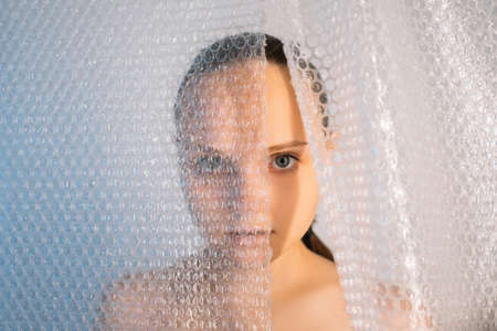 Covid-19 self isolation. Quarantine anxiety. Protective measures. Art portrait of disturbed woman hiding alone behind wrap drop curtain on light background.の写真素材