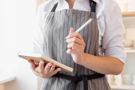 Female chef. Culinary recipe. Food cooking. Homemade cuisine. Unrecognizable woman in gray apron making notes in notebook light kitchen interior background.の写真素材