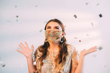 Pandemic celebration. Quarantine party. Festive surprise. Amused excited woman in gold chain face mask matching cocktail dress enjoying special day in confetti rain on pastel pink background.の写真素材