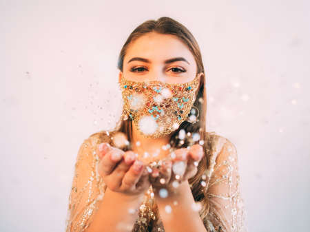 Pandemic Christmas. Festive greeting. Quarantine party celebration. Amused woman in glamour gold chain face mask catching bokeh light snowflakes in open palms on pastel pink copy space background.の写真素材