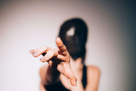 Woman protection. Female rights. Social isolation. Art portrait of suffering woman with head wrapped in black bag reaching out hand asking for help in spotlight on blur background.の写真素材