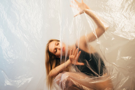 Art portrait. Woman protection. Social isolation. Help support. Insecure woman in black bodysuit hiding behind wrinkled polyethylene film drop on light copy space background.の写真素材