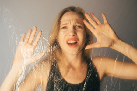 Female abuse. Defocused portrait. Woman protection. Social isolation. Frightened hurt lady looking for help through transparent creased texture polyethylene film on gray background out of focus.の写真素材