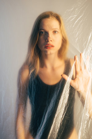 Pandemic self isolation. Defocused portrait. Coronavirus lockdown. Safety measures. Worried trapped woman looking through clear creased texture polyethylene film on gray background out of focus.の写真素材