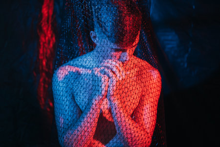 Man anxiety. Emotional crisis. Sorrow grief. Art portrait of depressed pensive shirtless guy praying in mesh trap in red blue neon light at night on blur dark background.の写真素材