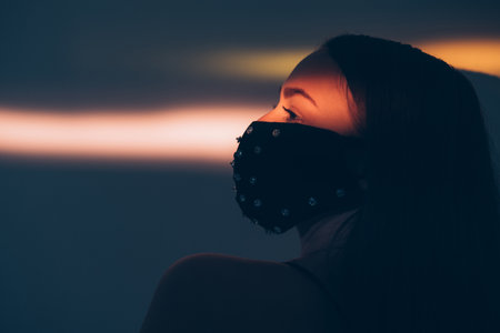 Pandemic fashion. Glamorous elegance. New normal style. Stay safe. Woman in black decorated crystal beads face mask posing on lights shadow side view copy space.の写真素材