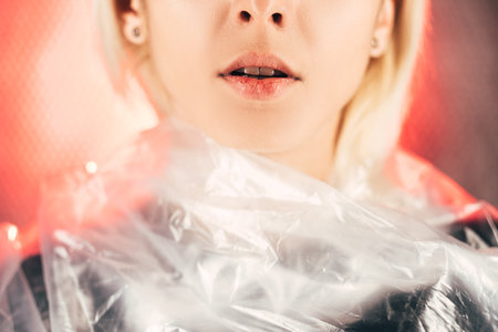 Female portrait. Plastic surgery. Face lifting. Lips contouring. Beauty procedure. Unrecognizable woman smooth skin wrapping clear cellophane film on peach red neon light background.の写真素材