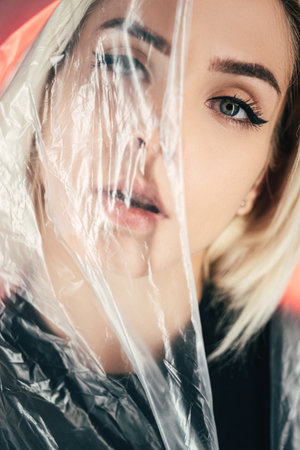 Art portrait. Plastic surgery. Fashion beauty. Face contouring. Woman smooth skin looking through transparent polyethylene film red neon light background.の写真素材