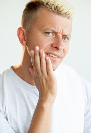 Male facial care. After shave skin moisturizing. Morning routine. Portrait of disturbed blond man in white t-shirt touching dry cheek isolated on light background.の写真素材