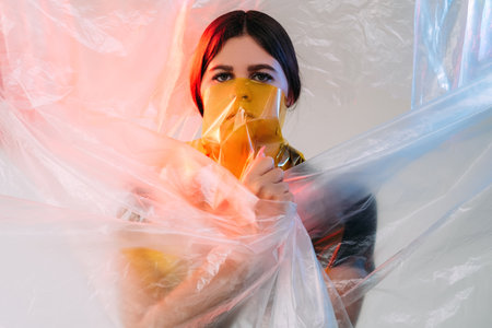 Pandemic self isolation. Covid-19 anxiety. Stay home. Lockdown ban. Art portrait of scared woman in yellow transparent face mask bag in pink blue neon light on wrapped in wrinkled polyethylene film.の写真素材