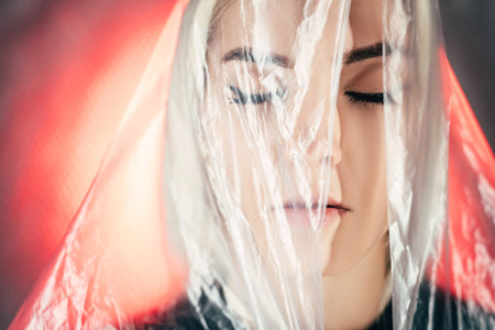 Art portrait. Waste reuse. Save planet. Self isolation. Environmental problems. Calm woman covered clear wrinkled polyethylene film closing eyes yellow red neon light background copy space.の写真素材